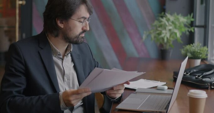 Middle Eastern entrepreneur reading business document and working with laptop typing in cafe. Businessman and paperwork concept.