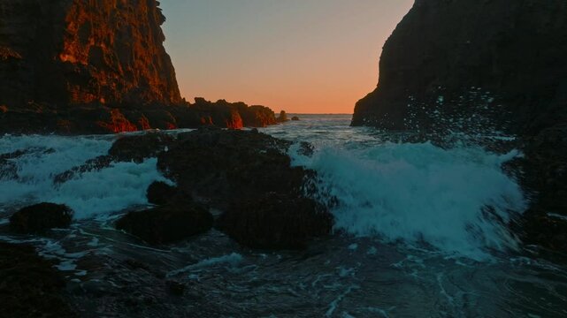 Ocean waves crashing through a cove over rocks and sand at sunset. Bethells Beach, Auckland, New Zealand.