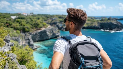 Fototapeta premium The photo shows a man standing with a backpack, overlooking the spectacular ocean cliffs and blue waters from a high vantage point on a sunny day with a few clouds in the sky.