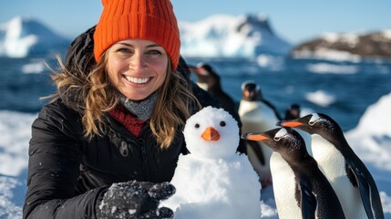 A joyful woman, wearing an orange beanie and black jacket, builds a snow penguin while surrounded by live penguins on a snowy landscape with glacial waters in the background.