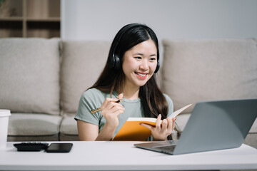 Smiling woman in headphones taking notes, motivated interested student studying online, using...