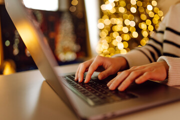 A person typing on a laptop in a cozy setting decorated with warm holiday lights during the festive...