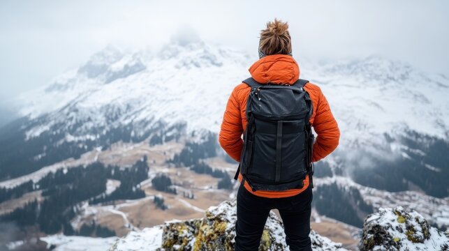A hiker wearing an orange jacket and a backpack stands on a snowy mountain ridge, gazing thoughtfully out over a picturesque valley, encapsulating the essence of wilderness exploration.