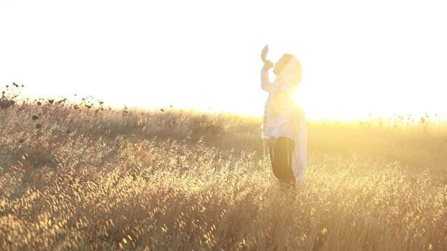 Jewish man in a traditional tallit prayer shawl blowing the ram's horn shofar, in the field against sunrise sky on Rosh HaShana and Yom Kippurim 