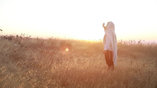 Jewish man in a traditional tallit prayer shawl blowing the ram's horn shofar, in the field against sunrise sky on Rosh HaShana and Yom Kippurim 