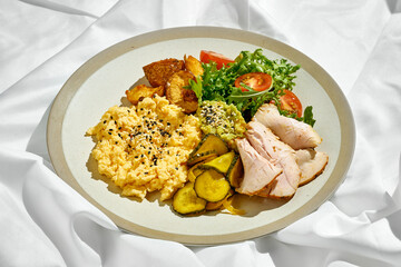 A balanced and healthy breakfast bowl filled with scrambled eggs, roasted potatoes, fresh salad, and sliced ham. The bowl is topped with avocado and sesame seeds.