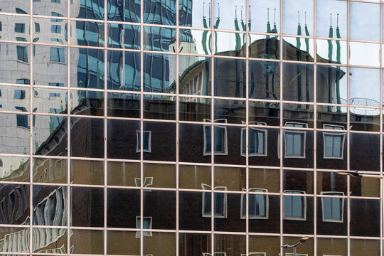 Numerous city buildings, including a historical structure with flags, are reflected on the glass facade of a modern skyscraper, symbolizing urban complexity and contrasts.