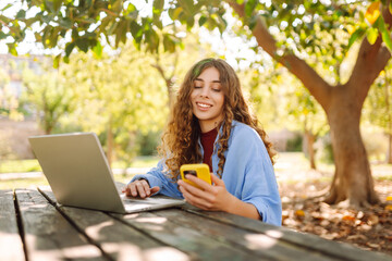 A young woman sits at a wooden table in a park, focused on her laptop while holding a smartphone. Education and modern technologies.