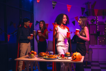 Asian friends, middle-aged man and young woman, enjoy Halloween office party with vampire costumes, fried chicken, grilled food, grape wine, champagne, RGB lights, festive decorations in the night.