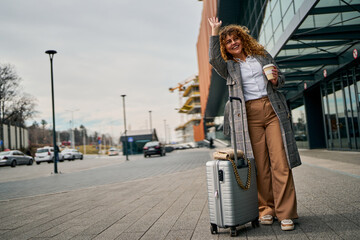 After the flight, a curly-haired redhead outside the airport raises her hand, signaling for a taxi with her suitcase.