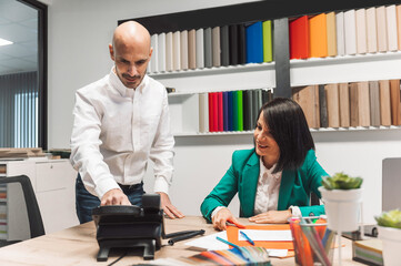 A professional meeting in an office setting. A man in a white shirt is showing something on a phone to a woman in a green blazer, who is smiling. The background features colorful materials and a moder