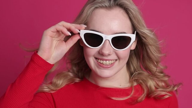 Smiling young caucasian blond woman with transparent ceramic braces on teeth isolated on pink backdrop. Girl takes off her stylish sunglasses and blinks at camera. Beauty, health and fashion concept.