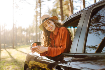 The young woman leans out of the car window, basking in the sunlight and enjoying the natural surroundings of a serene forest road. The concept of active lifestyle, travel, tourism.