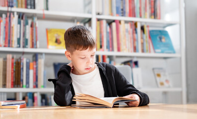 11-year-old teenage boy in a white T-shirt reads an interesting book in the library against the...