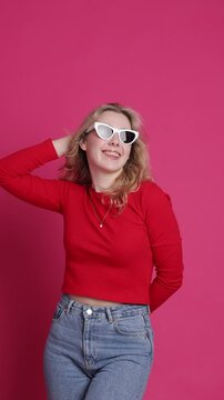 Cheerful young caucasian woman with transparent ceramic braces on teeth isolated on pink backdrop. Blond girl in eyeglasses looks at camera. Windy effect with fan. Beauty, health and fashion concept.
