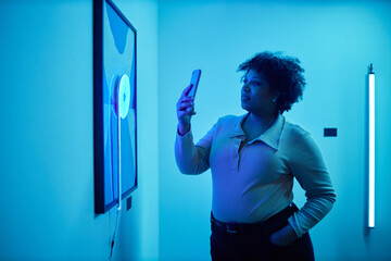 Side view portrait of African American woman looking at paintings in blue neon light during modern...