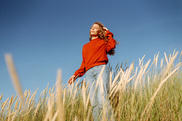 Young woman enjoying a sunny day outdoors in tall grass while wearing a cozy red sweater....