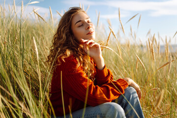 Young woman enjoying a sunny day outdoors in tall grass while wearing a cozy red sweater....