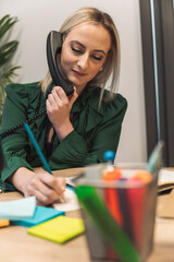 A woman in a green blouse is sitting at a desk, talking on a vintage telephone while writing notes. A colorful container with stationery items is visible in the foreground.