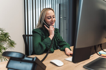 A woman in a green blouse sitting at a desk, talking on the phone while working on a computer. The office has a modern design with a plant in the background.