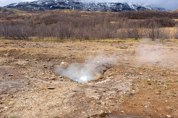 A misty geothermal landscape at Geysir, Iceland. Steam rises from numerous hot springs, creating a surreal and ethereal atmosphere. The volcanic activity is evident in the bubbling mud pools.