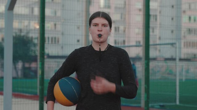 Young Caucasian woman poses confidently in a basketball uniform, holding a ball and whistling. Basketball judge or referee at the match in stadium. Determination and athletic power. Student at college