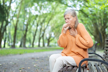 Senior woman sitting on park bench struggles with breathing. Image captures concern and health issues in tranquil natural environment. Depicts aging, respiratory problems, need for medical attention.