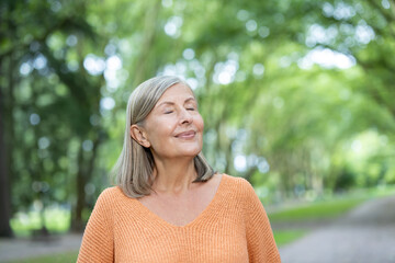 Elderly woman with short gray hair wearing orange sweater stands with eyes closed, enjoying...