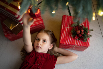 Little girl lies on floor reaching up to touch bright ornament hanging from Christmas tree. Vibrant presents adorned with red polka dots and striped paper surrounding child