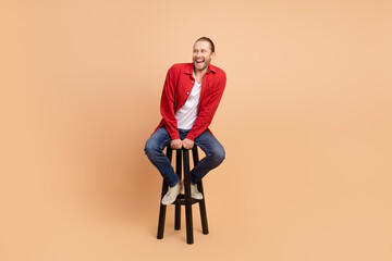 Full size photo of nice young man sit stool look empty space wear red shirt isolated on beige color...