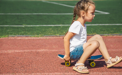 Modern little girl sitting on skateboard in the sport field. Active kids. Sports. Entertainment.