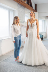 Naklejka premium A bridal designer fitting a wedding dress on a smiling bride in a bright, airy studio. The dress features intricate lace details and a flowing skirt, while the designer looks pleased with the fit.