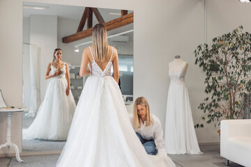 A bride trying on a wedding dress in a boutique, with a consultant adjusting the gown. The scene features a large mirror reflecting the bride and elegant dresses in the background.