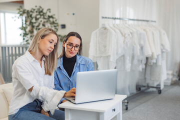 Obraz premium Two women collaborating on a laptop in a bright, modern workspace. One woman has long blonde hair and wears a white shirt, while the other has brown hair and glasses, dressed in a blue striped shirt.
