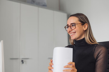 A young woman with glasses smiles while holding a stack of papers in an office setting. She is wearing a black turtleneck and appears engaged and happy, with a modern workspace in the background.