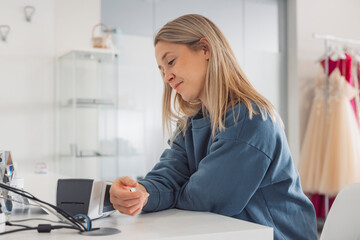 A young woman with long blonde hair sitting at a white table, looking thoughtfully at a small device. The background features a modern interior with a display case and dresses hanging.