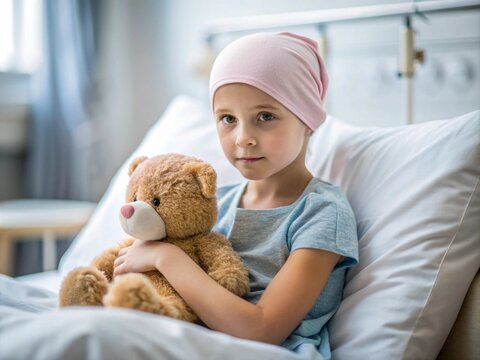 A little girl with cancer sits on a bed with a teddy bear in a hospital room.