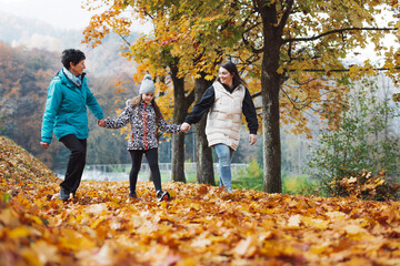 Naklejka premium A family walking together on a path covered with autumn leaves. The scene features two adults and a child, all dressed in colorful jackets, enjoying a sunny day in a park surrounded by trees with vibr