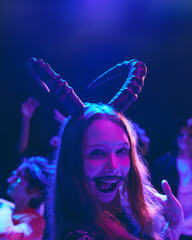 Portrait of smiling young girl with creepy makeup and animal horns on head celebrating Halloween with friends at nightclub. Concept of Halloween, holiday, party, leisure