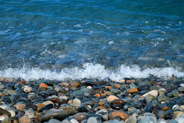 Foamy strong sea transparent waves crashing on pebble beach on Black Sea coast. Waves splashing on stones shore in sunny weather. Clear wate in sea waves broken on pebble colorful shore.