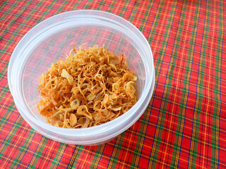 Deep-fried savory crispy sliced shallots, stored in a transparent plastic container, placed on a vibrant plaid red and green tablecloth, creating a colorful and traditional food presentation