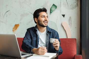 Young indian freelancer smiling and looking away while working on laptop in cafe