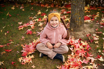 A smiling girl sits cross-legged on the grass, surrounded by vibrant red and yellow autumn leaves while enjoying a beautiful day outdoors in a park.