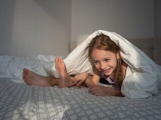 A cunning little girl lies on her stomach on the bed, looks out from under the duvet, next to her...