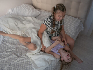 The older sister is sitting on the bed and tickling the younger one lying in front of her. A little...