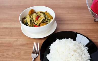 Steamed rice with a fork on the side and bamboo shoot curry with green leafy vegetables, pumpkin and chilli. In a white cup with a saucer In the upper right corner is fruit on a wide wooden table.
