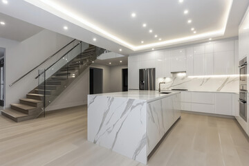 Modern kitchen interior with white marble island, sleek cabinets, and built-in appliances under recessed lighting. Glass staircase with metal railing in contemporary open-plan space. Luxury home