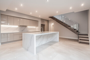 Sleek modern basement kitchen featuring a large white marble island, glossy cabinets, and a glass staircase with ambient lighting