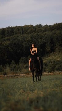 Young woman rides bareback on a black horse in a lush green field, surrounded by dense forest. She is dressed in a black strapless outfit and sits gracefully on the horse, exuding calm and confidence
