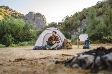 Fototapeta premium Adult caucasian man sit in front tent and use laptop on camping trip